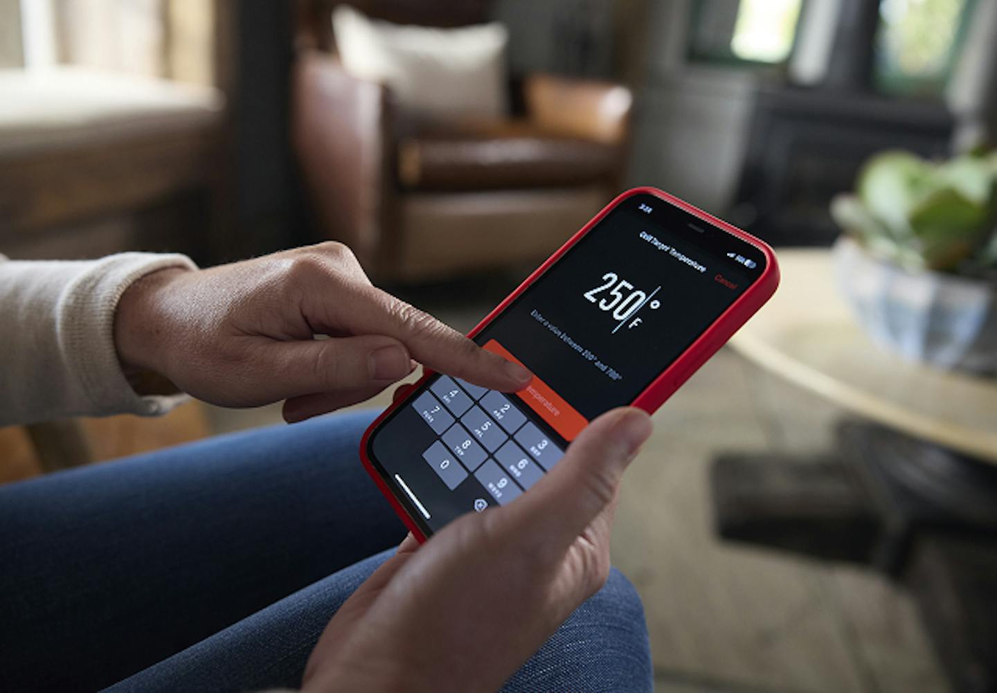 A person adjusts temperature settings on a smartphone displaying 250&deg;F in a cozy indoor environment.
