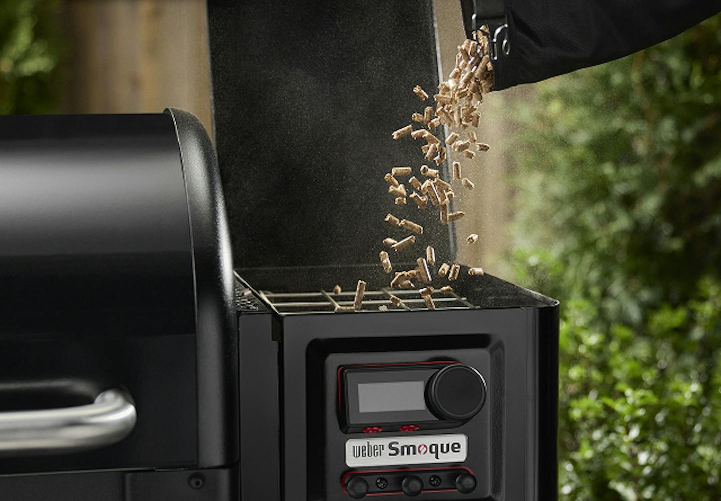 A hand pours wood pellets into the hopper of a Weber Smoque grill, surrounded by greenery.