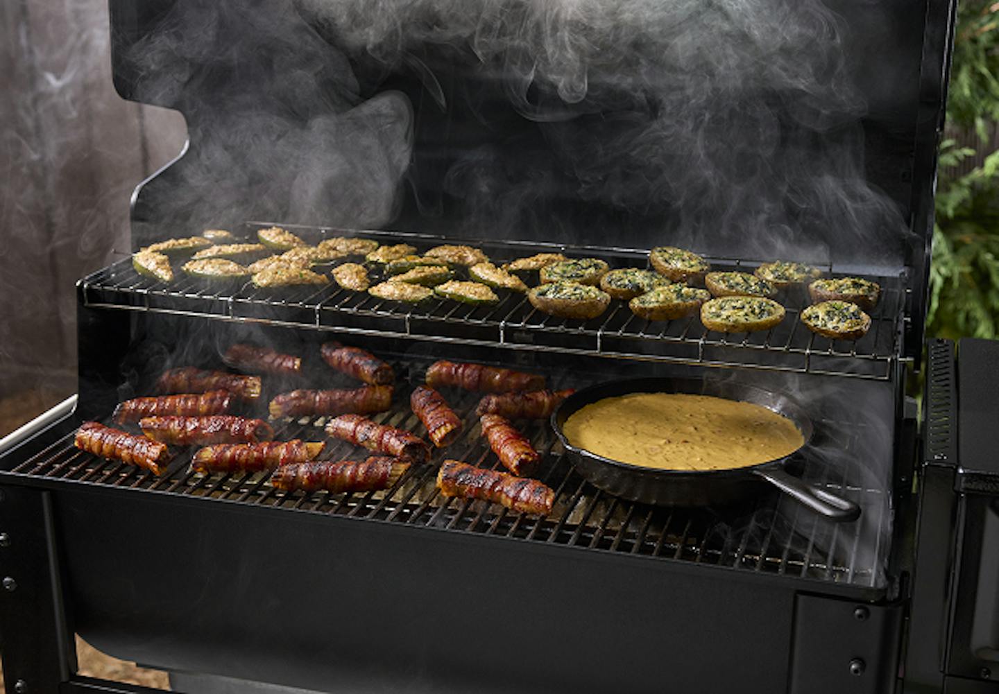 A smoky barbecue scene featuring grilled sausages, stuffed mushrooms, and a pan of sauce on a black grill.