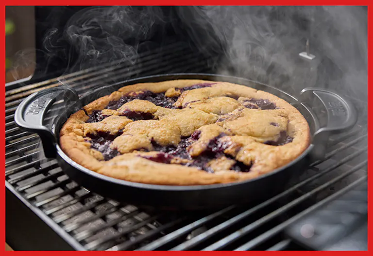 A bubbling berry pie baked on a grill, emitting steam, with a golden-brown crust in a black cast iron pan.
