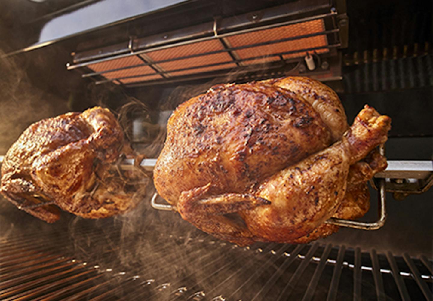 Two whole chickens cooking on a rotating rotisserie and smoke rising from the grill