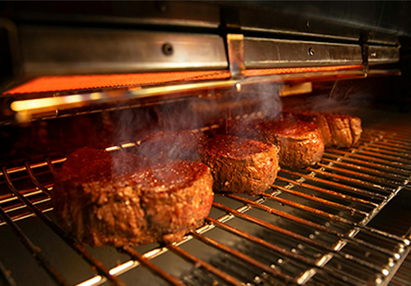 Four steaks grilling on a heated grill and smoke rising from the steaks