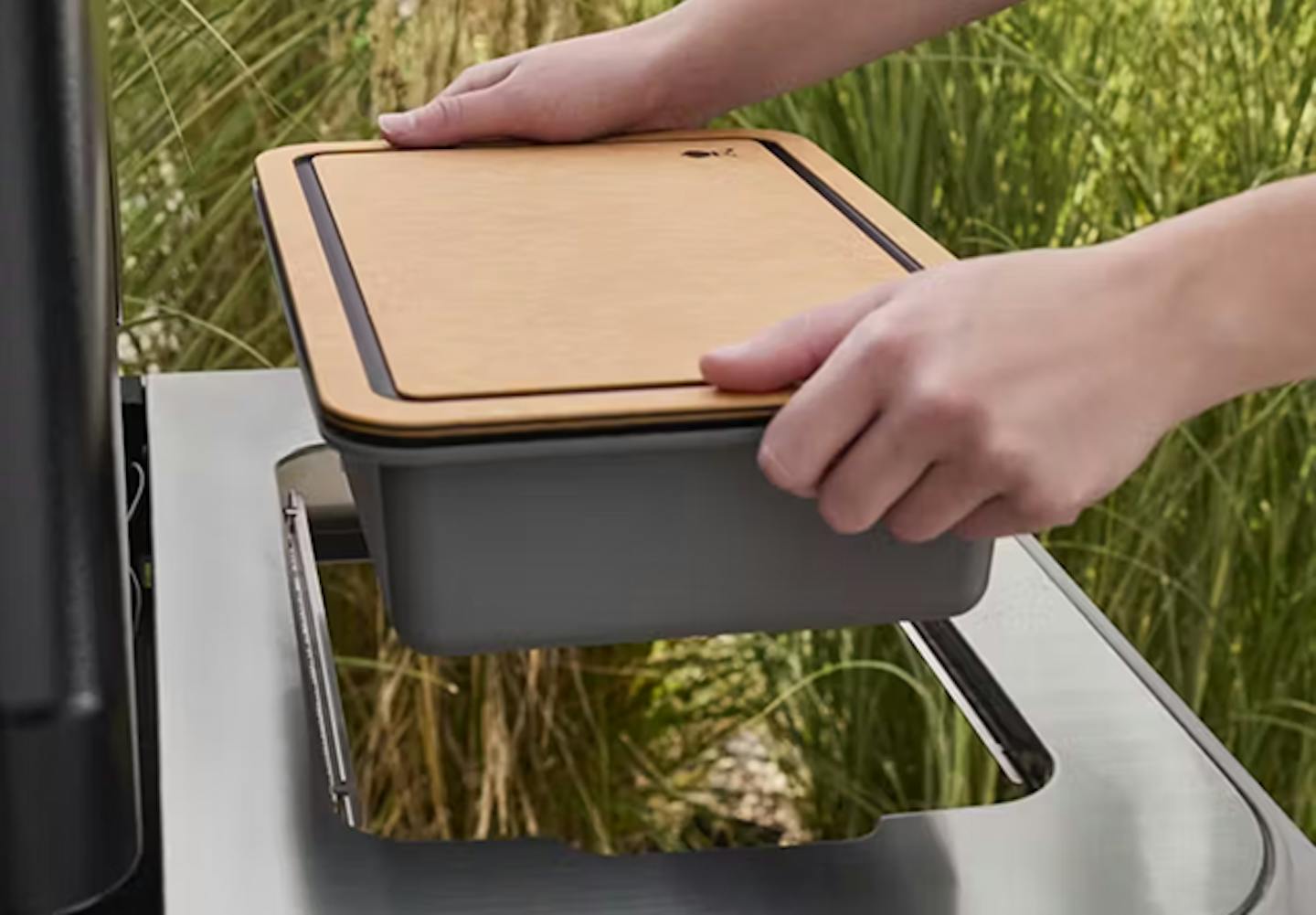 En person plasserer et trelokk på en grå beholder mens han setter den inn i en elegant utegrill, omgitt av grønne planter.