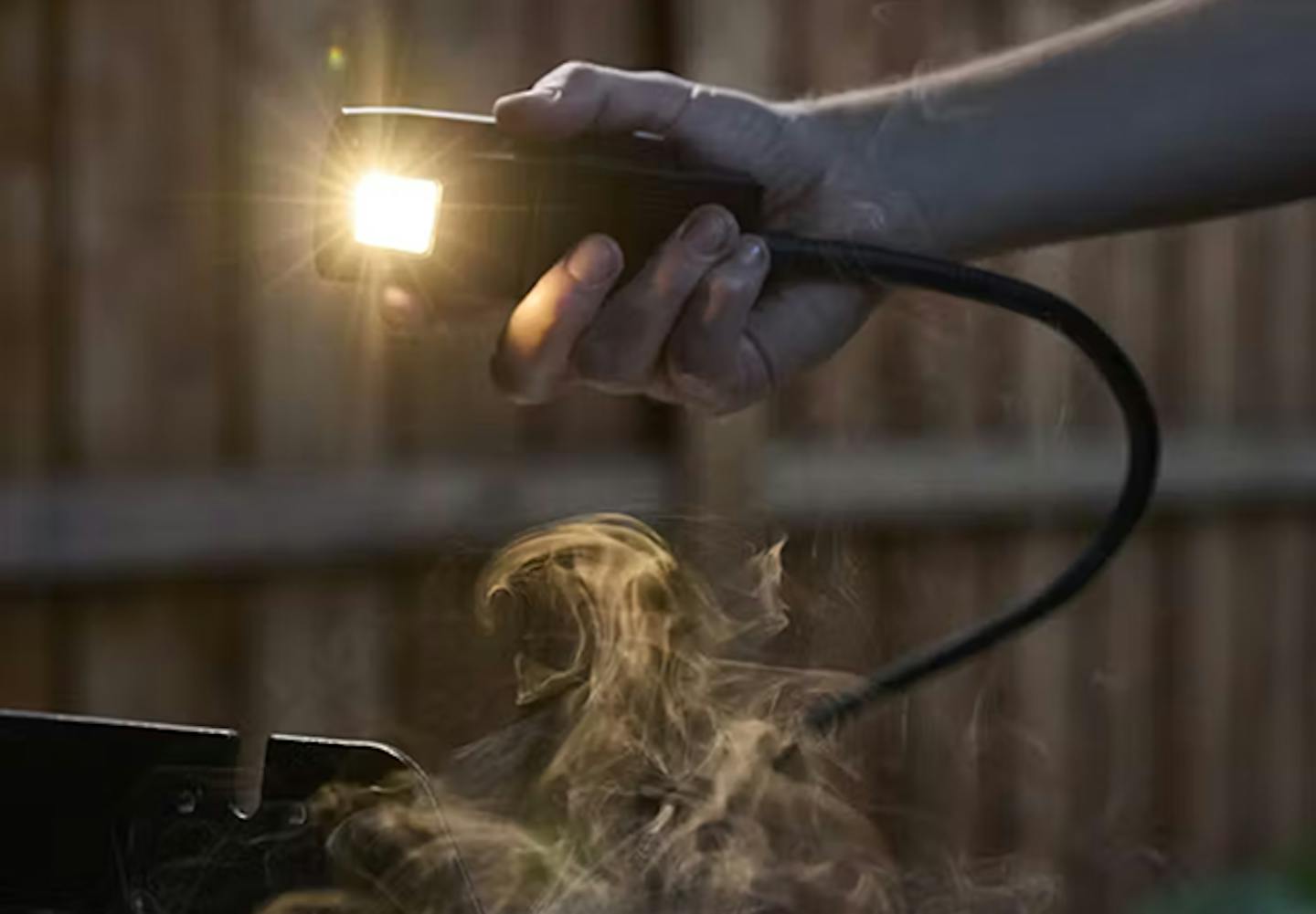 En hånd holder en bærbar lampe som lyser sterkt, omgitt av virvlende røyk, i et svakt opplyst utendørs miljø.