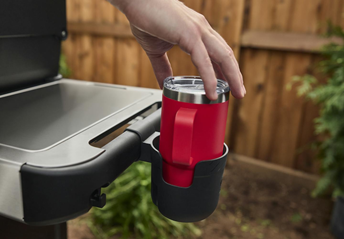 En hånd plasserer et rødt drikkebeger i en koppholder på en grill, med et tregjerde og grøntområder i bakgrunnen.